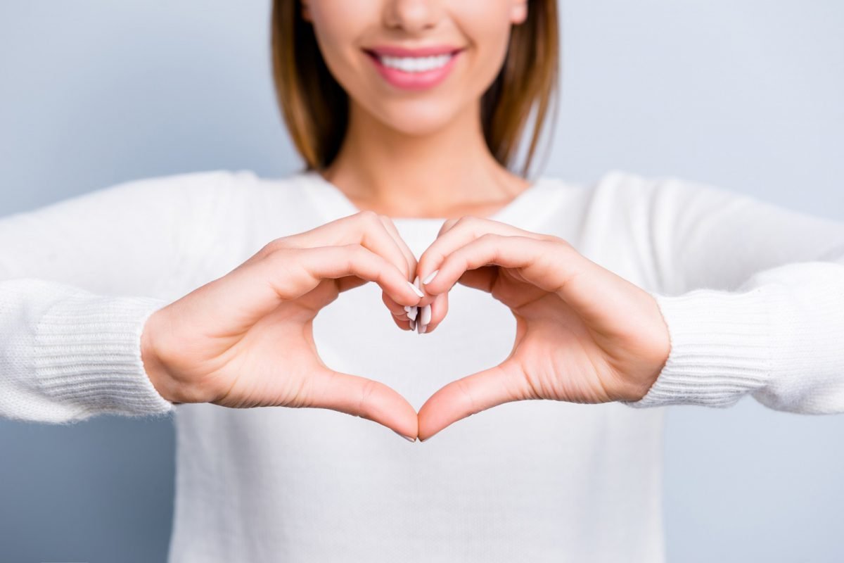Cropped portrait of smiling woman in white outfit showing focused love symbol heart figure with fingers isolated on grey background
