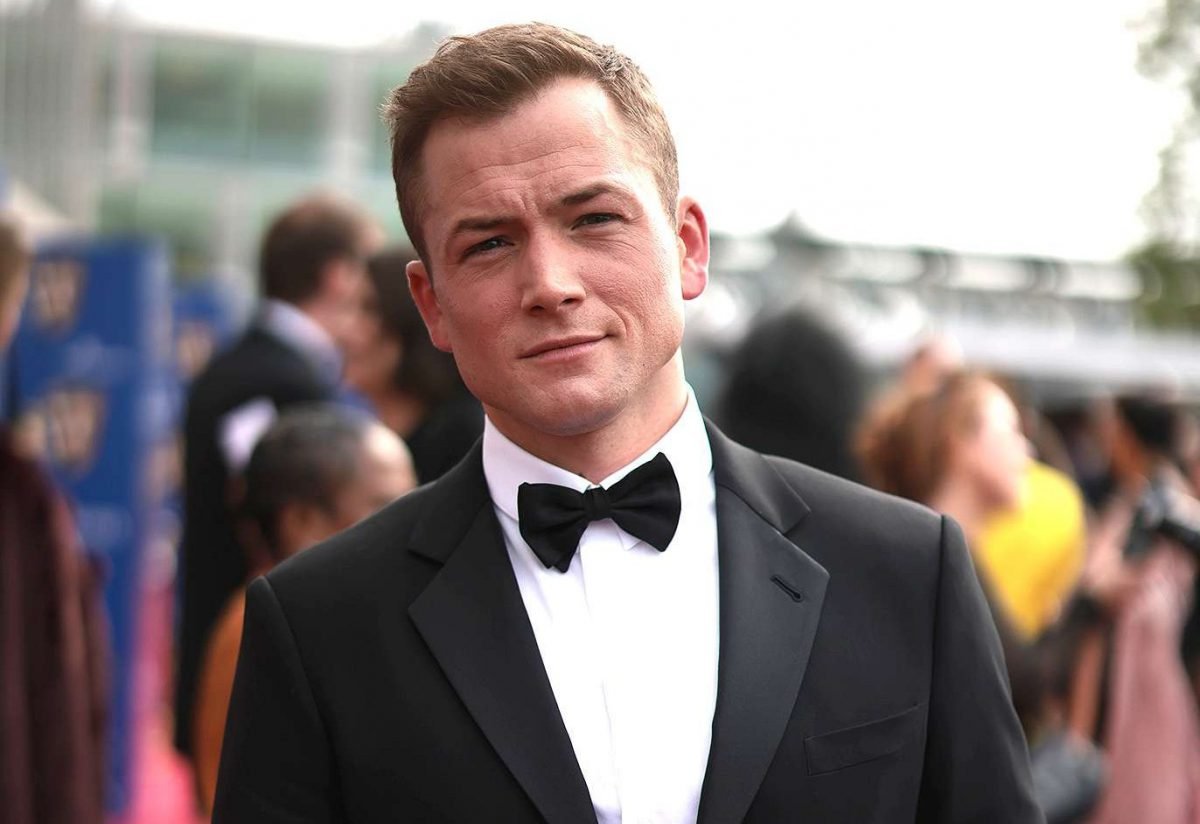 Taron Egerton, Forehead, Hairstyle, Bow tie, Dress shirt, Coat, Beard, Gesture